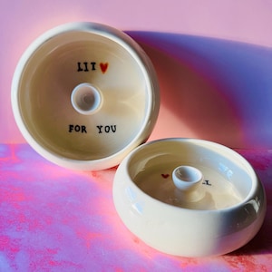 May include: Two off-white ceramic bowls with a glossy finish. The bowls have a circular shape with a small, raised center. One bowl has the text "LIT" with a heart and "FOR YOU". The other bowl has a heart and the letter "I".