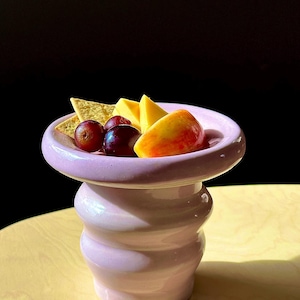 Puede incluir: Un cuenco de cerámica rosa con un borde ancho y una base ondulada y texturizada. El cuenco está lleno de un pequeño aperitivo de uvas, cubos de queso y galletas.