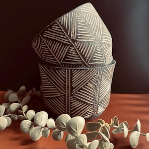 Puede incluir: Dos cuencos de cerámica marrón oscuro apilados con un diseño geométrico tallado en color marrón claro. Los cuencos son cilíndricos con una parte superior ligeramente ensanchada. Una ramita de eucalipto artificial está en primer plano.