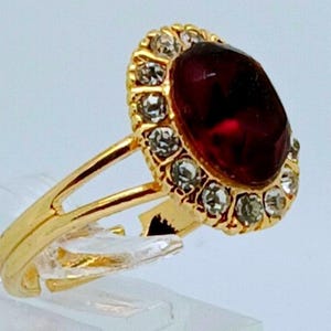 May include: A gold-colored ring featuring a large, oval, deep red gemstone surrounded by a halo of smaller, clear stones. The ring's band is split and has a delicate design, showcasing a classic jewelry style.