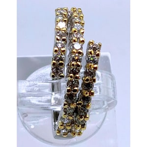 May include: A three-band ring with a spiral design. The ring features a silver band with gold accents and is encrusted with small, clear and brown gemstones. The ring is displayed on a clear, plastic stand.