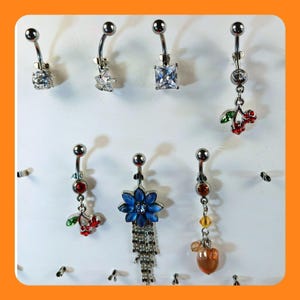 May include: A collection of seven belly button rings displayed on a white surface. The rings feature various designs, including clear and colored gemstones, floral patterns, and dangling charms. The jewelry is made of silver-colored metal.