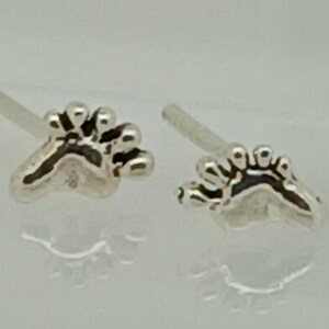 May include: A pair of silver-colored stud earrings shaped like tiny footprints. Each earring features a detailed paw print design with visible toe pads and claws. The earrings are set against a light background.