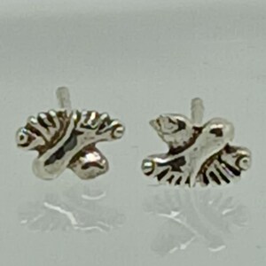 May include: A pair of small, silver-colored stud earrings. Each earring features a detailed, stylized design resembling a bird or angel with outstretched wings. The earrings have a reflective surface and are set against a light background.