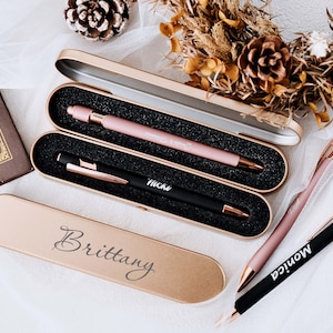 May include: A selection of personalised pens in gold cases. One case is open, showing a pink pen and a black pen. The pens have names engraved on them. A case is engraved with the name "Brittany".
