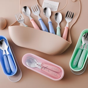 May include: A collection of baby feeding utensils in various colors. The set includes spoons and forks with pastel-colored handles, stored in matching cases. A beige bib and wooden ball are also present.
