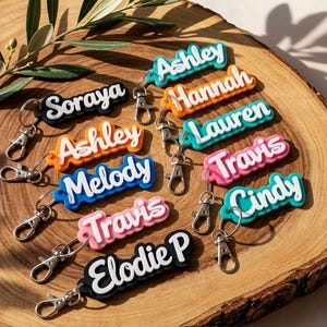 Peut inclure: Plusieurs porte-clés nominatifs colorés avec des fermoirs argentés. Les noms incluent "Ashley", "Hannah", "Lauren", "Travis", "Cindy", "Melody", "Elodie P" et "Soraya". Chaque porte-clés a un schéma de couleurs différent.