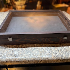 May include: A dark stained wooden display case with a glass top. The case has a rectangular shape with a hinged lid and metal clasps. The interior has a dark gray surface.
