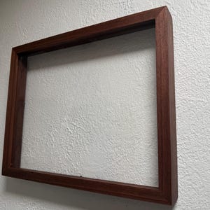 May include: A rectangular, dark brown wooden picture frame. The frame is empty, with a visible grain pattern. The frame is mounted on a textured white wall. The frame's dimensions are not visible.