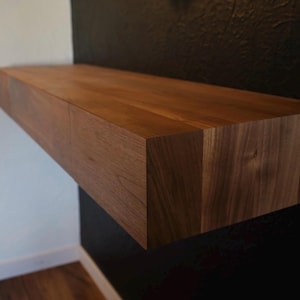 May include: A dark brown wooden floating shelf with a simple design. The shelf is mounted to a black wall and has a smooth, natural finish.