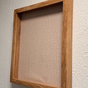 Op de afbeelding: Een vierkante houten fotolijst met een lichtbruine, getextureerde stoffen achterkant. De lijst is gemaakt van lichtgekleurd hout en is gemonteerd op een witte, getextureerde muur. De lijst is leeg.