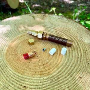 May include: A brass pen with a dark brown leather grip featuring cross-stitch detailing. The set includes a hexagonal brass cap, a small metal component, two white cylindrical inserts, and a bullet-shaped tip with a red plastic cover.