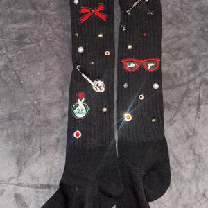 Peut inclure: Chaussettes noires montantes avec divers embellissements brodés colorés, dont un nœud rouge, une paire de lunettes rouges avec les mots "hello you" dessus, un geste de la main vert et une épingle à nourrice argentée.