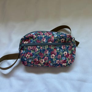 May include: A floral print fanny pack with a dark blue background and a pattern of pink, red, and blue flowers. The bag has a brown strap and a zipper closure. The bag is approximately 10 inches wide.