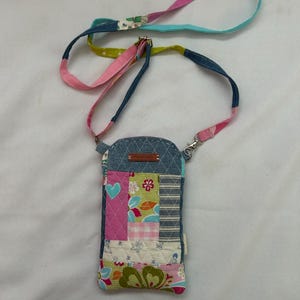 May include: A patchwork cell phone crossbody bag with a denim top and a colorful strap. The bag features a variety of patterned fabrics in pink, blue, green, and white. The strap is made of multiple colors and patterns.