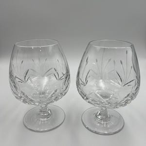 May include: Two clear crystal brandy snifters. Each glass features a rounded bowl, a short stem, and a wide, flat base. The bowls are etched with a delicate floral design. The glasses are displayed against a plain, light background.