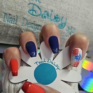 May include: Close-up of a hand with red, blue, and white nail art. The nails are long and have a matte finish. The hand is holding a white flower with a blue center and the words "Teal Daisy" written on it.