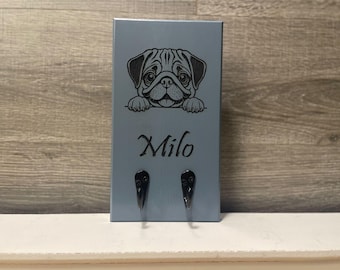 May include: A light blue key holder featuring a pug illustration and the name "Milo" in script. The key holder has two black hooks for hanging keys. The pug is depicted in black, with its paws resting on the top edge.