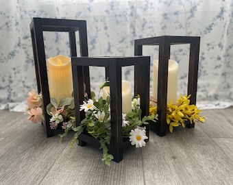May include: Three black wooden candle lanterns with faux flowers. The lanterns are square and have a simple design. The flowers are white and yellow.