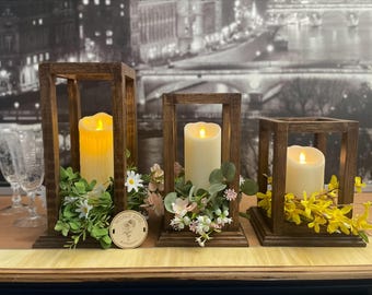 May include: Three wooden candle lanterns with faux candles and floral arrangements. The lanterns are different sizes and are arranged in a row on a wooden surface. The flowers are white, pink, and yellow.