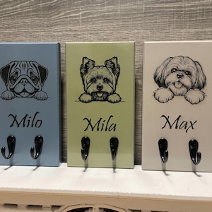 May include: Three rectangular wooden key holders with dog illustrations. Each has a different dog breed, name, and two black hooks. The colors are light blue, light green, and off-white. The names are Milo, Mila, and Max.