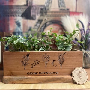 May include: A wooden planter box with a natural wood finish. The planter has an engraved design of flowers, butterflies, and the text "Grow with Love". The planter is sitting on a wooden base and has a small wooden tag with the text "Moonlit Rose Workshop".