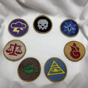 May include: A collection of embroidered patches with various symbols. The patches are circular with gold-colored borders. Symbols include a skull, lightning bolt, scales, fire, a compass rose, a tank, and an eye within a triangle.