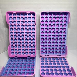 May include: Two rectangular storage organizers featuring a grid of circular holes. Each unit has a pink exterior frame and a light purple interior base. The design includes a vertical back section and a horizontal bottom section with rounded edges.