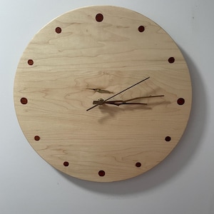 May include: A round wooden clock with a light wood grain finish. The clock face features dark red dots marking the hours and thin, dark metal hands. The clock is mounted on a white wall.
