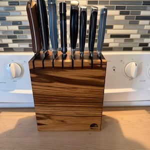 May include: A wooden knife block with a set of knives. The block is made of light brown wood with a natural grain pattern. The knives have dark handles and silver blades. The block is sitting on a light-colored wooden surface.
