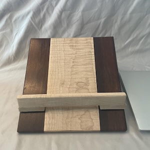 May include: A wooden laptop stand with a light brown top and dark brown sides. The stand is made of two pieces of wood that are joined together with a wooden bar.