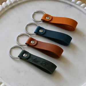 May include: Four leather keyrings in orange, teal, brown, and dark green. Each keyring has a silver metal ring and a matching rivet. The dark green keyring is engraved with the name "Alex B."