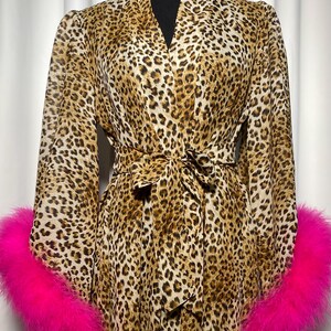 May include: A leopard print robe with long sleeves and a matching tie belt. The cuffs are trimmed with bright pink faux fur. The robe is displayed on a black mannequin.