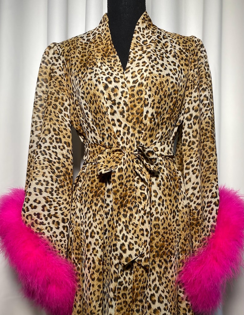 May include: A leopard print robe with long sleeves and a matching tie belt. The cuffs are trimmed with bright pink faux fur. The robe is displayed on a black mannequin.