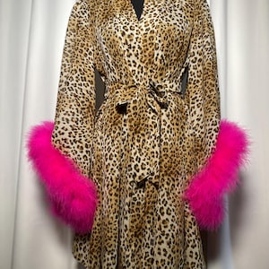 May include: A leopard print robe with long sleeves and a matching belt. The robe has bright pink feather trim on the cuffs. The garment is displayed on a black mannequin against a white backdrop.