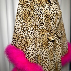 May include: A leopard print dressing gown with vibrant pink feather trim on the sleeves. The gown features a tie waist and is displayed on a black mannequin. The fabric has a brown and black spotted pattern.