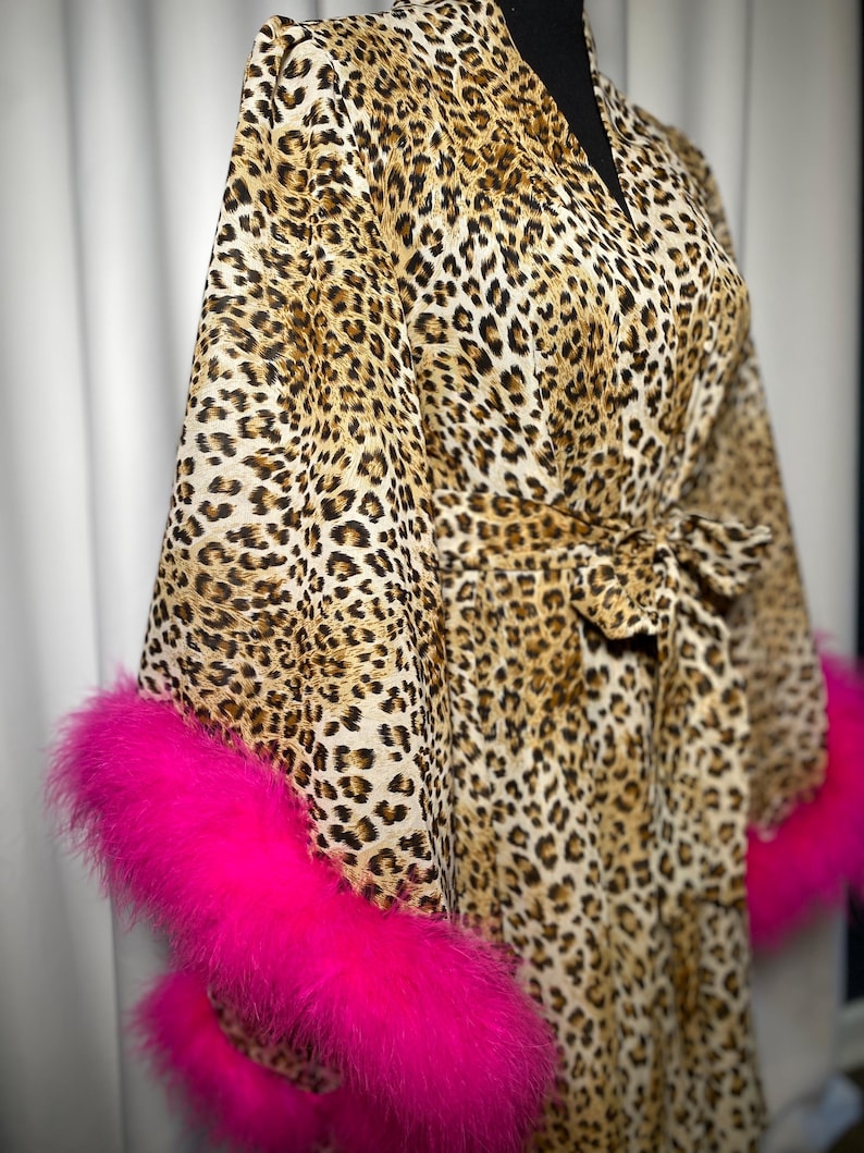 May include: A leopard print dressing gown with vibrant pink feather trim on the sleeves. The gown features a tie waist and is displayed on a black mannequin. The fabric has a brown and black spotted pattern.