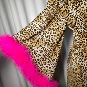 May include: A leopard print dress with long sleeves and a bright pink feather trim on the cuffs. The dress features a brown and tan spotted pattern. The sleeves are flared, and the fabric appears lightweight.