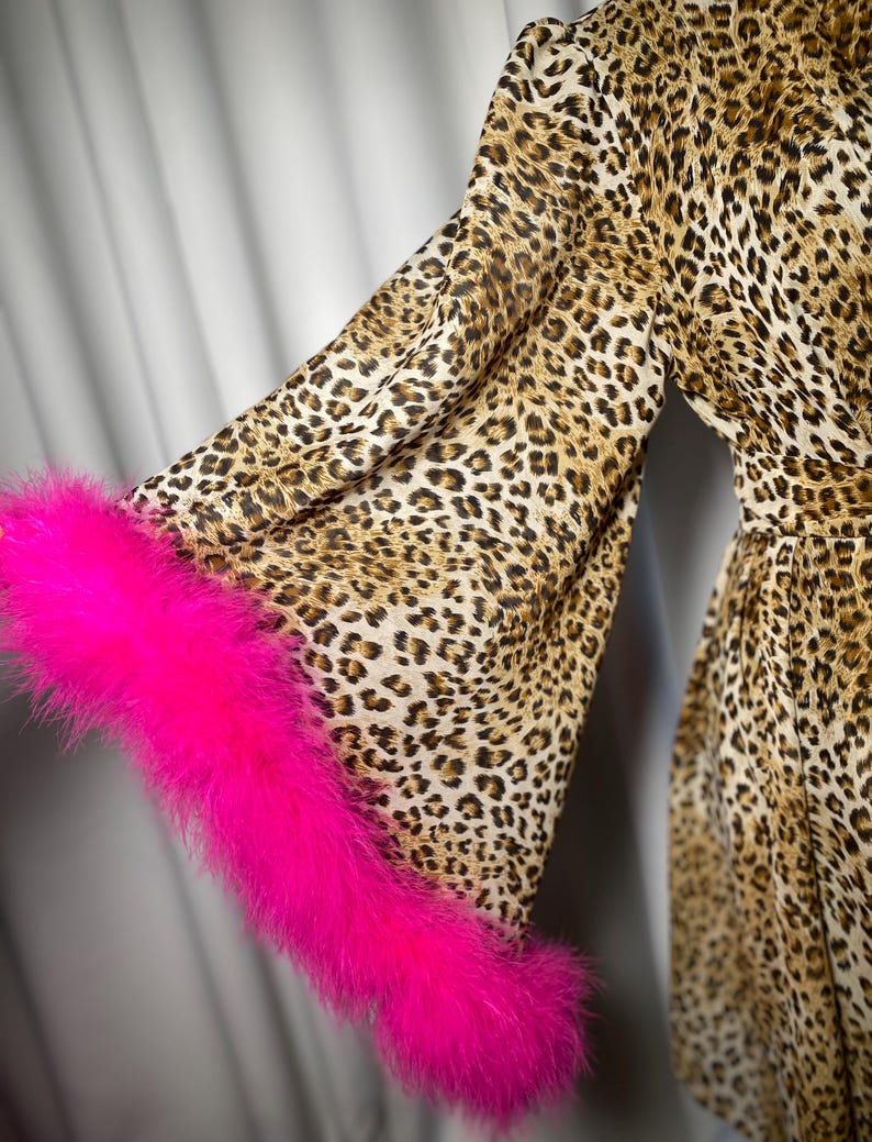 May include: A leopard print dress with long sleeves and a bright pink feather trim on the cuffs. The dress features a brown and tan spotted pattern. The sleeves are flared, and the fabric appears lightweight.