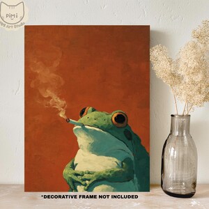 Op de afbeelding: Een rechthoekige wandkunstprint met een groene kikker met grote oranje ogen die een brandende sigaret vasthoudt met zichtbare rook tegen een effen, gebrande oranje achtergrond. De tekst onderaan luidt DECORATIVE FRAME NOT INCLUDED.
