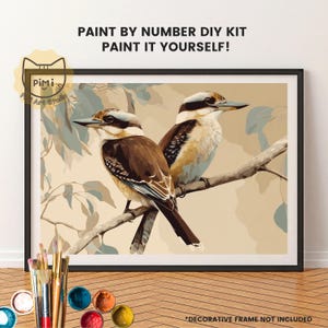 May include: A paint by number DIY kit featuring two kookaburra birds perched on a branch. The text reads PAINT BY NUMBER DIY KIT PAINT IT YOURSELF. The artwork displays brown, cream, and muted blue tones. Includes a set of paintbrushes and paint pots.