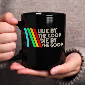 May include: Black ceramic mug with the text "LIVE BY THE GOOP DIE BY THE GOOP" in white. The mug features a colorful design of blue, green, yellow, and red stripes. The mug is held by a person.