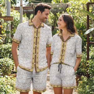 May include: Matching white pajama sets with a repeating cartoon bear pattern. The sets include a button-down shirt and shorts, trimmed with brown and gold accents. The couple is standing in a garden setting.