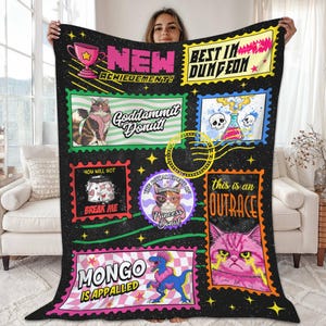 May include: Black fleece blanket with colorful postage stamp-style graphics. The blanket features various cat illustrations, text like "NEW ACHIEVEMENT!" and "BEST IN DUNGEON", and a dinosaur. The blanket is held up by a person.