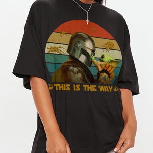 A person wearing a black t-shirt with a graphic design featuring a character from the Star Wars franchise, specifically The Mandalorian, holding a small green alien-like creature.