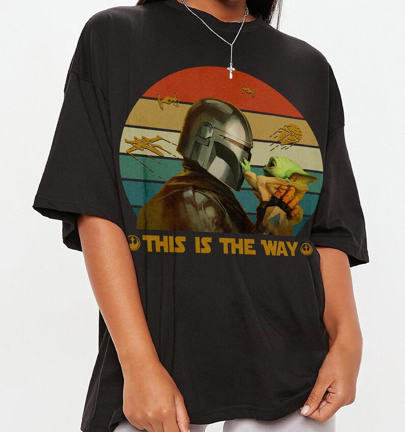 A person wearing a black t-shirt with a graphic design featuring a character from the Star Wars franchise, specifically The Mandalorian, holding a small green alien-like creature.