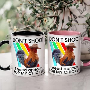 May include: Two white ceramic mugs with black and pink interiors. Each mug features a rooster wearing a hat, with text that reads "DON'T SHOOT" and "I need apricots FOR MY CHICKEN". The mugs have black and pink handles.