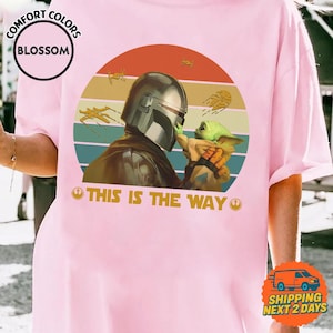 a person wearing a pink t-shirt with a graphic design depicting a character holding a baby, along with the text "THIS IS THE WAY" and a Star Wars logo.