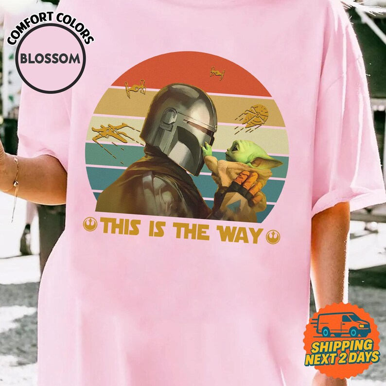 a person wearing a pink t-shirt with a graphic design depicting a character holding a baby, along with the text "THIS IS THE WAY" and a Star Wars logo.
