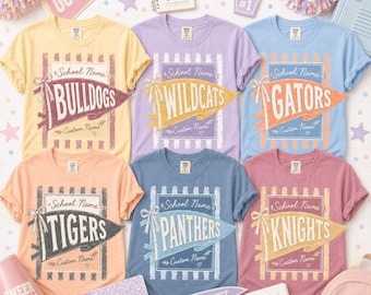 May include: Six short-sleeve crew neck t-shirts in yellow, lavender, light blue, peach, slate blue, and mauve. Each shirt features a central graphic of a pennant flag with school mascot names: Bulldogs, Wildcats, Gators, Tigers, Panthers, and Knights.